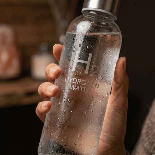 Dłoń trzymająca elegancką szklaną butelkę z napisem H2 Hydrogen Water, pokrytą kropelkami wody.